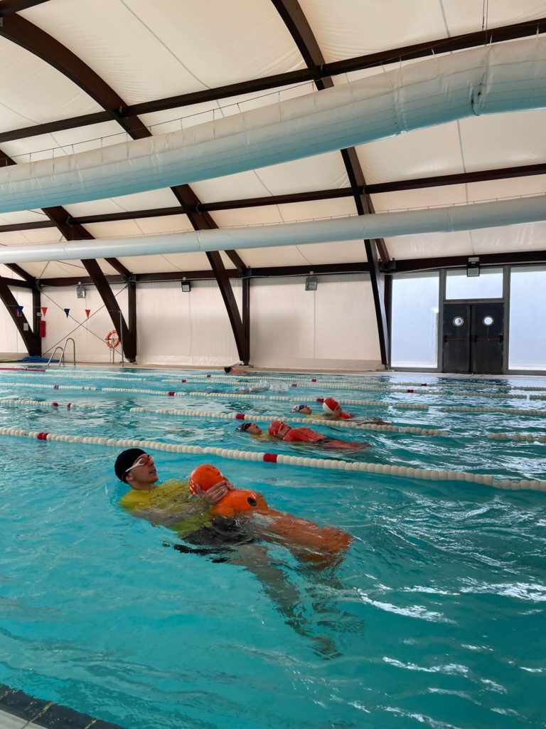 Grupo de alumnos en una piscina realizando entrenamiento de socorrismo acuático, remolcando muñecos de salvamento